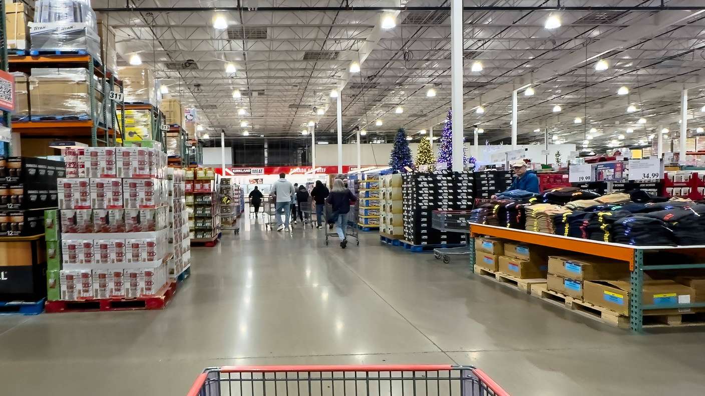 Blick hinter den Einkaufswagen auf den Gang eines Costco-Großhandelsclubs mit Massenware, Bekleidungstischen und einem geschmückten Weihnachtsbaum während des geschäftigen Weihnachtsgeschäfts.
