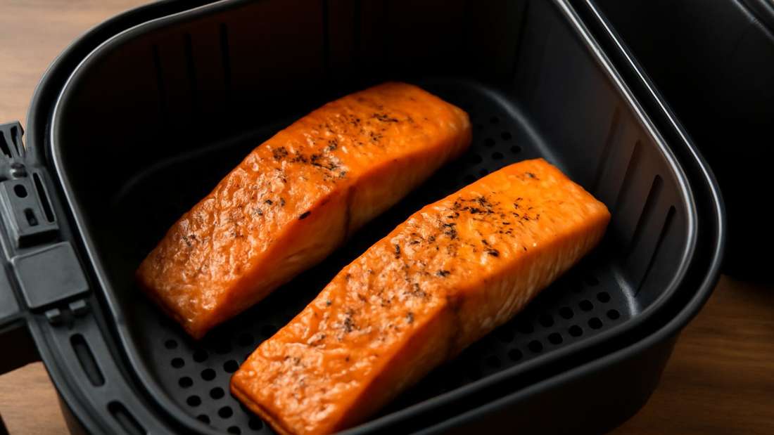 Zwei Lachsfilets im Airfryer, die in einem Airfryer liegen. Die Filets haben eine goldbraune Kruste und sind mit schwarzem Pfeffer gewürzt. Der Hintergrund ist weich und verschwommen, wobei der Fokus auf den Lachsfilets liegt, die in einem schwarzen Airfryer zubereitet werden.