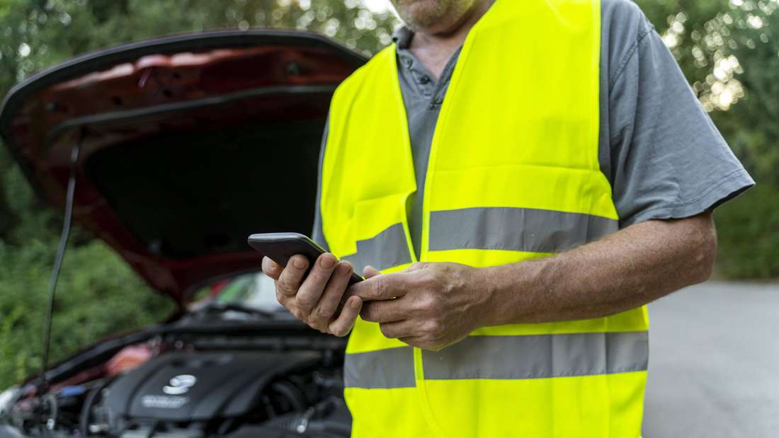 Ein Mann in einer gelben Warnweste steht vor einem roten Auto mit offener Motorhaube.