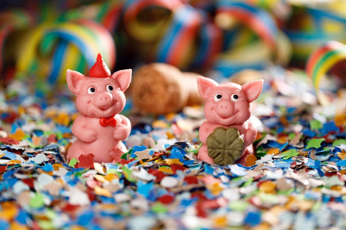 Zwei Marzipanschweine sitzen auf einem Meer aus Konfetti. Das eine davon hält noch ein vierblättriges Kleeblatt. 