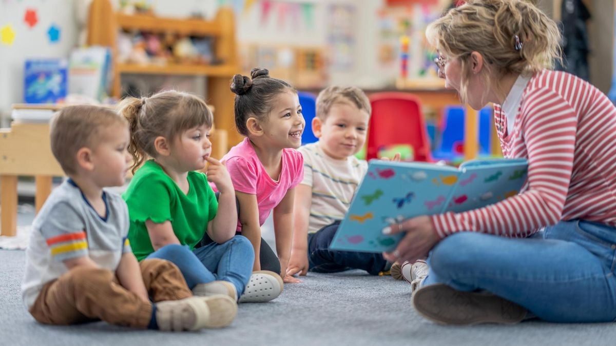 A preschool teacher sits on the floor of her classroom with a small group of students as she reads them a book. The children are each dressed casually and are focused on the story. Reading is Fun!