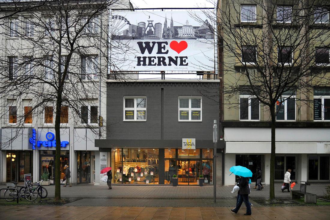 Eine urbane Straßenszene in Herne mit mehreren Gebäuden, wobei ein graues Geschäftshaus in der Mitte ein großes weißes Werbeschild mit der Aufschrift „WE LVOE HERNE“ trägt, während im Vordergrund eine Person mit einem türkisfarbenen Regenschirm vorbeigeht und kahle Bäume sowie weitere Geschäfte die winterliche Atmosphäre der Fußgängerzone prägen.