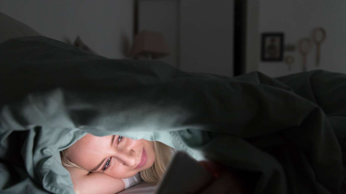 Fröhliche junge Frau im Bett liegend unter der Decke mit Handy in der späten Nacht, kann nicht schlafen
