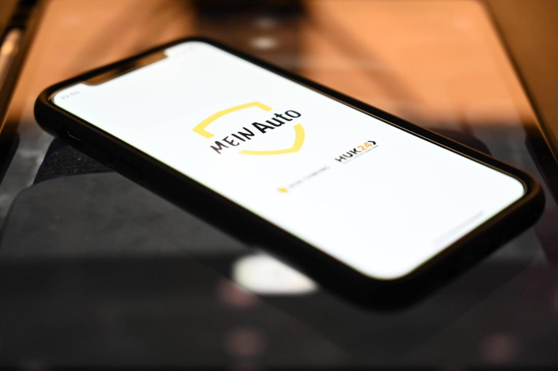 Ein schwarzes Smartphone, das auf einer dunklen Oberfläche liegt und auf dessen Bildschirm eine weiße App-Oberfläche mit dem gelben Logo „MEIN AUTO“ und darunter einem kleineren schwarzen Symbol zu sehen ist.