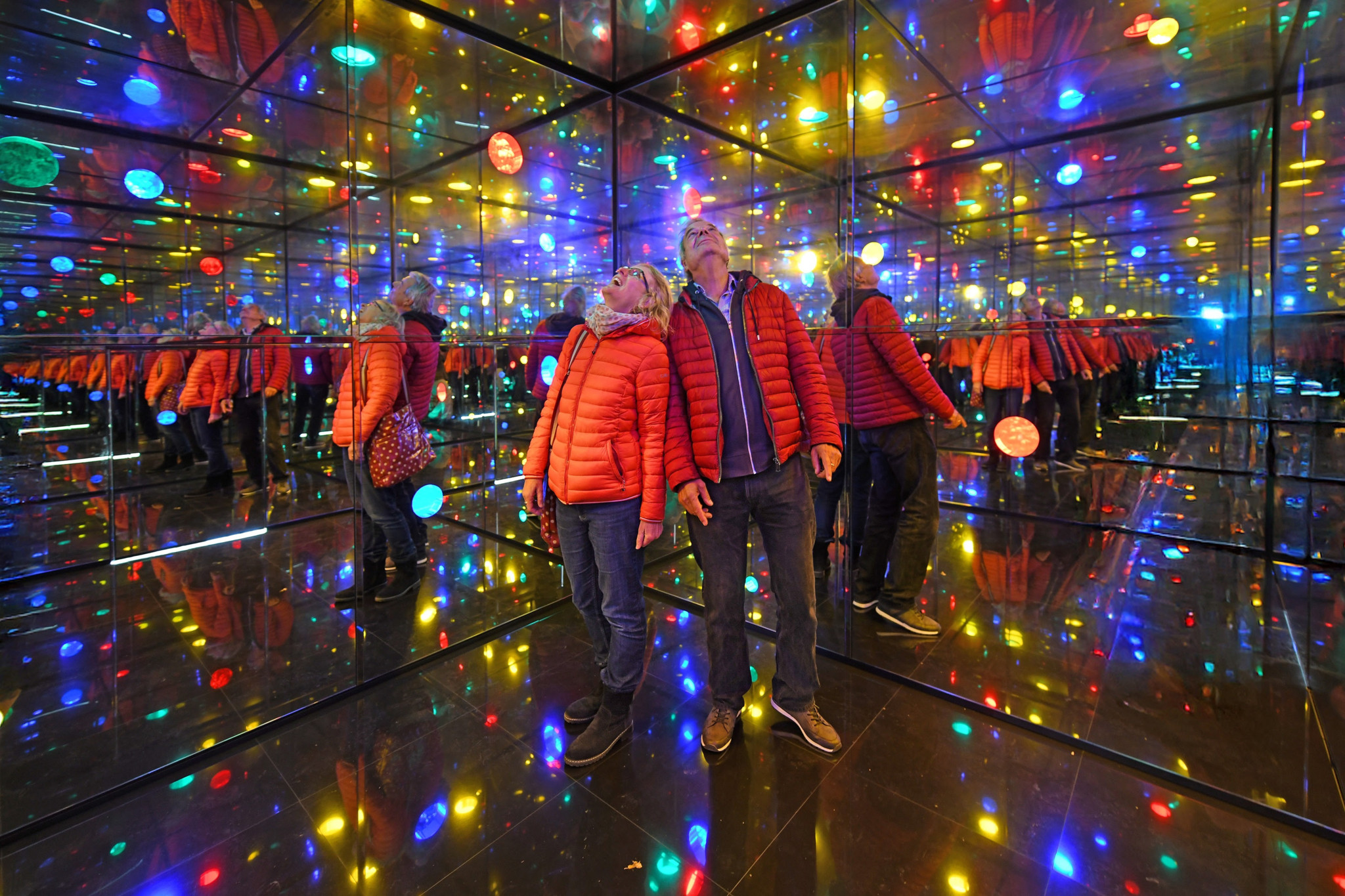 Personen in roten Jacken stehen in einem spiegelnden Raum mit bunten Lichtern, die eine Illusion von Unendlichkeit schaffen.