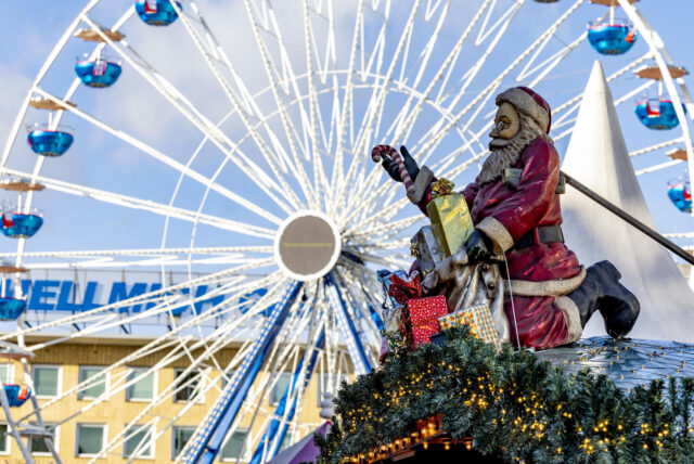 Eine Santa-Figur sitzt auf einem Dach, umgeben von Weihnachtsdekoration, während im Hintergrund ein Riesenrad für einen Weihnachtsmarkt steht.