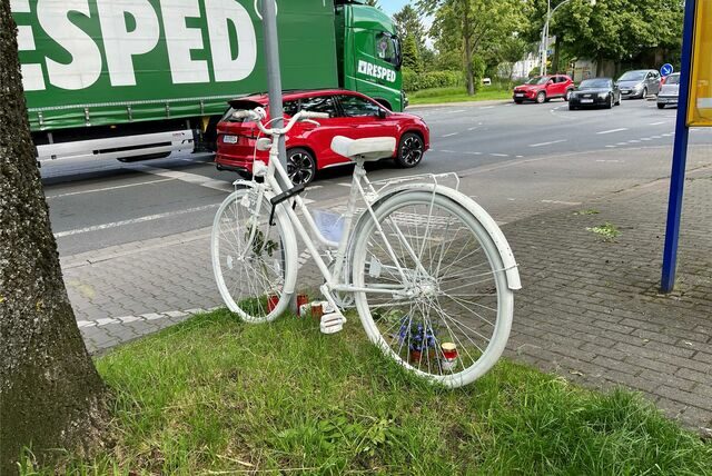 Ein weißes Fahrrad steht am Rande der Wiese, in der Nähe von parkenden Autos und einem Supermarkt.