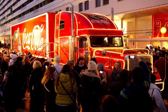 Ein roter Lkw mit beleuchteter Coca-Cola-Werbung steht unterhalb eines Gebäudes, während Menschen Fotos machen und sich versammeln.
