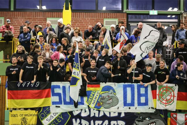 Eine Gruppe von Fans steht in einer Sporthalle, sie hält Fahnen und Banner und feuert ihr Team an.
