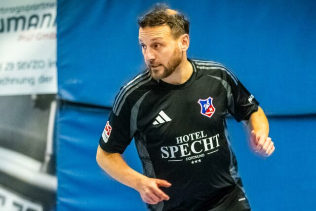 Ein sportlicher Mann in einem schwarzen Trikot mit Sponsorenlogos läuft in einer Sporthalle und konzentriert sich auf das Spiel.