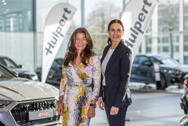 Zwei Frauen stehen in einem Autohaus, lächelnd, vor einer Auswahl von Fahrzeugen. Im Hintergrund sind Fahnen mit dem Schriftzug "Hülpert".