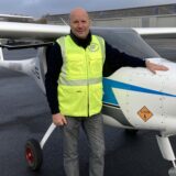 Ein Pilot steht neben einem einmötigen Propellerflugzeug. Im Hintergrund sind geschlossene Hangars zu sehen.