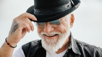 Ein lächelnder Mann mit weißem Bart, Sonnenbrille und schwarzem Hut, der seinen Hut leicht anhebt.