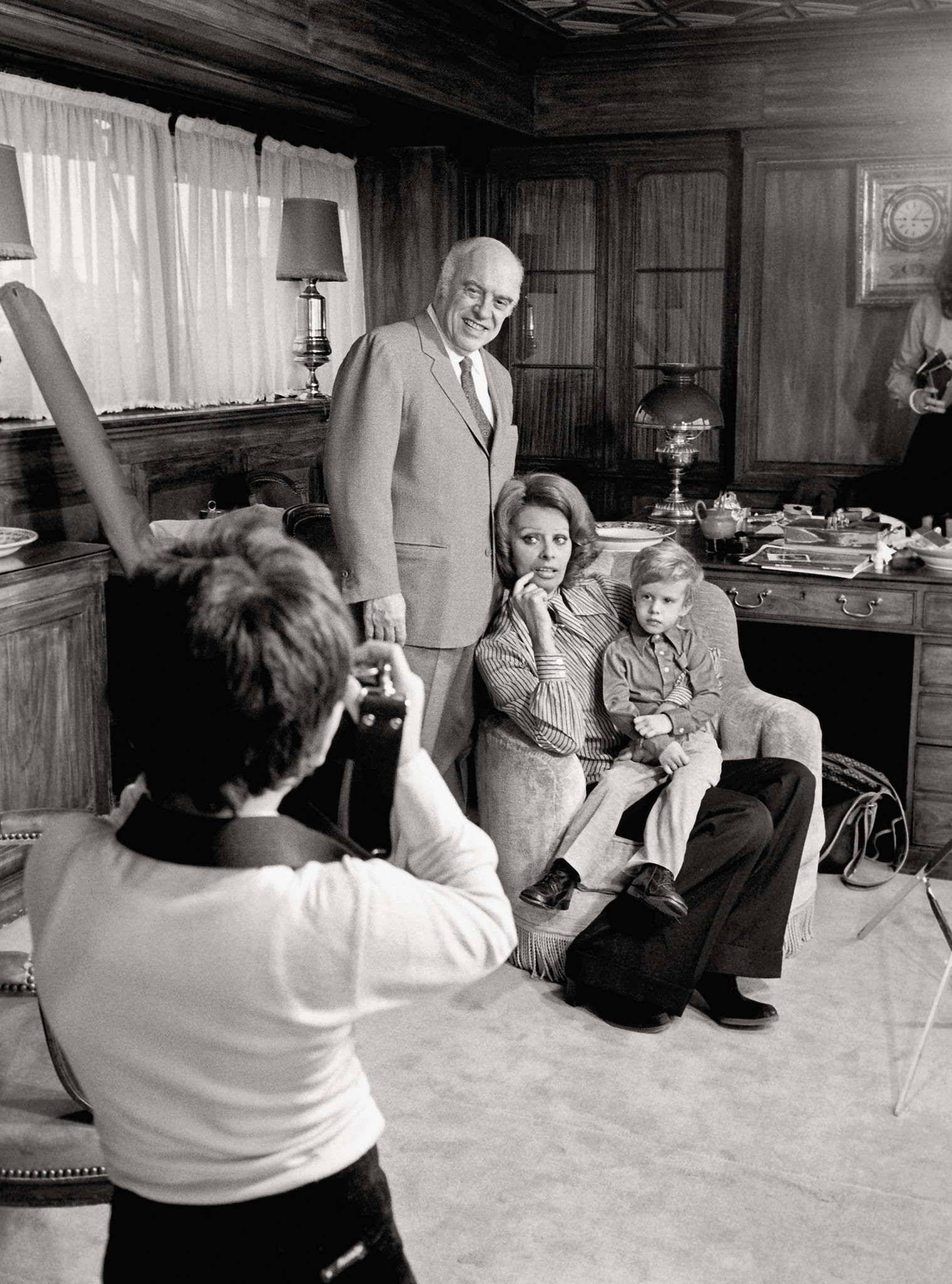 Eine Familie posiert für ein Foto in einem elegant eingerichteten Raum. Ein Mann steht hinter einer Frau, die auf einem Sessel sitzt, und ein Kind sitzt neben ihr. Ein Fotograf im Vordergrund macht das Bild.