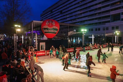 Menschen aller Altersgruppen fahren Schlittschuh auf der festlich beleuchteten Eisbahn, umringt von Weihnachtsschmuck und einer großen Feldschlößchen-Werbung.