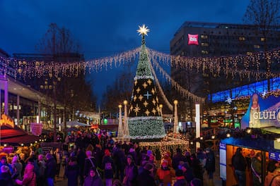 Der beleuchtete Weihnachtsbaum und funkelnde Lichterketten auf der Prager Straße erhellen den Winterabend, während Besucher die warme Atmosphäre des Marktes genießen.
