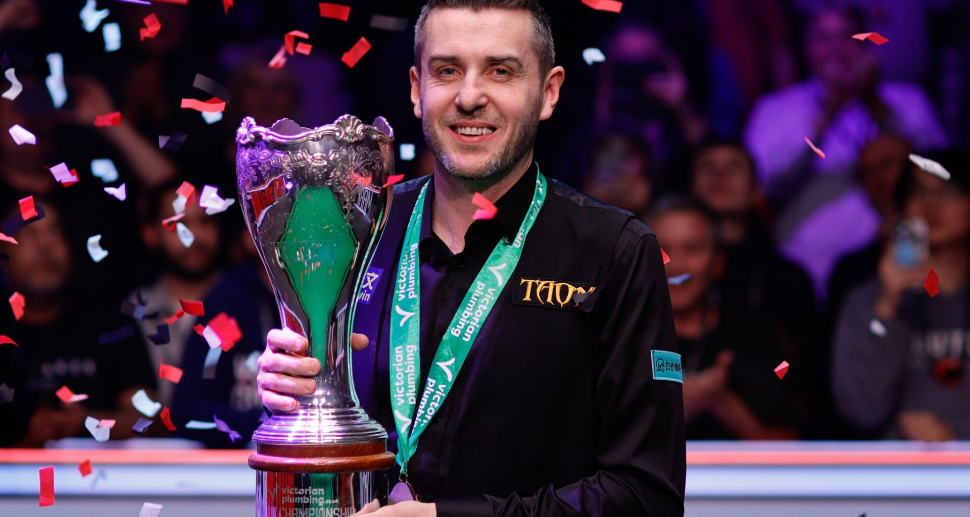 Mark Selby hält lächelnd die Trophäe der UK Championship, während Konfetti auf ihn herunterregnet.