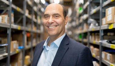 Mann in Anzug lächelnd in Lagerregal mit Kartons und Produkten.