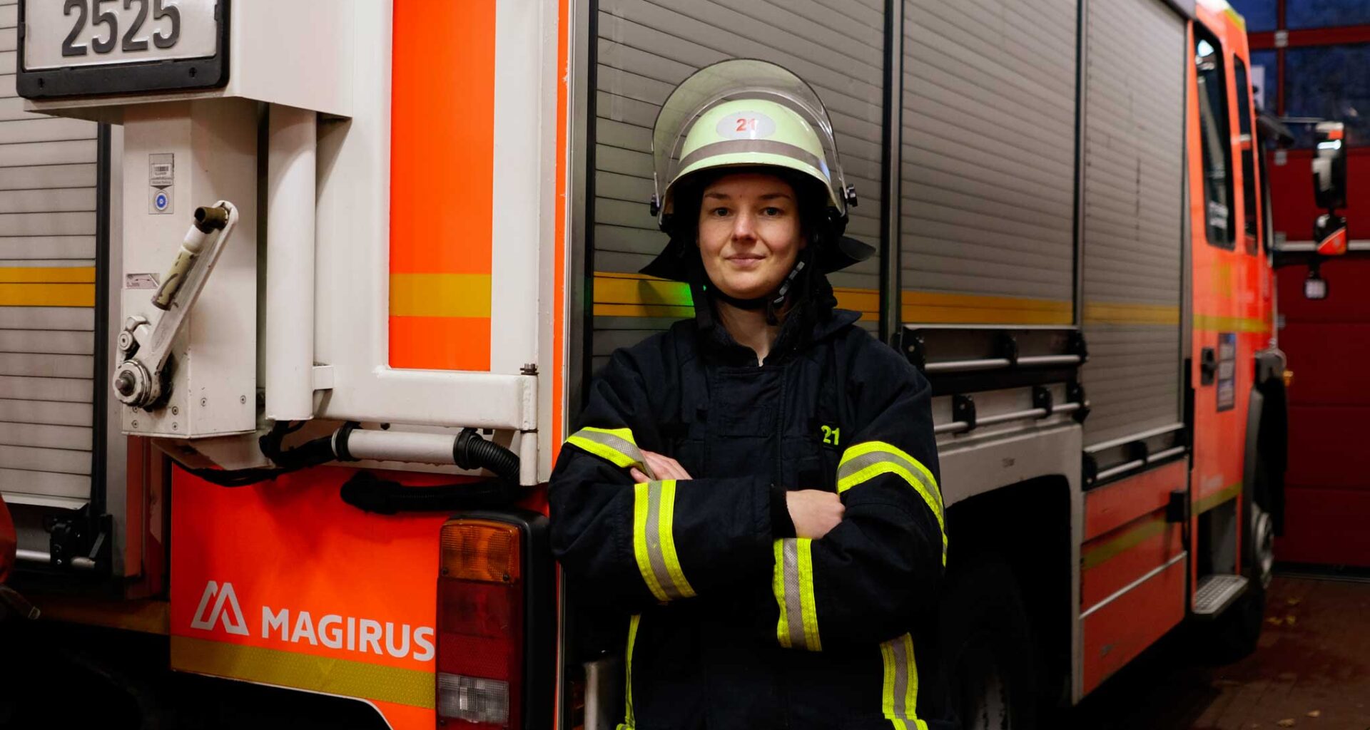 Feuerwehrfrau Nora Tenbusch steht vor einem Einsatzwagen der Feuerwehr. Sie trägt die Schutzkleidung der Feuerwehr und hat einen Helm auf dem kopf. Sie schaut in die Kamera und hat ihre Arme verschränkt.
