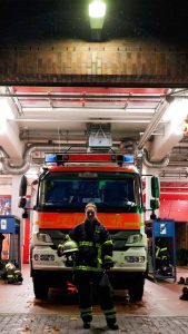 Nora Tenbusch steht vor einem Einsatzwagen der Feuerwehr. Sie hat sich einen Helm unter den Arm geklemmt und schaut in die Kamera.