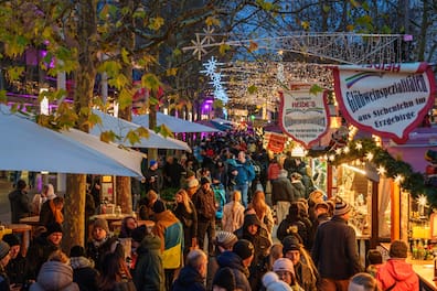 Dicht gedrängte Besucher schlendern entlang der geschmückten Marktstände, mit Tannenbäumen und funkelnden Lichterketten im Hintergrund.