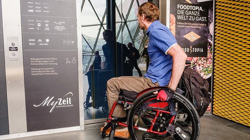 Bildbeschreibung: Jemand mit einem Rollstuhl vor einem Gebäude.