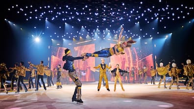 Eiskunstläufer hebt Partnerin bei HOLIDAY ON ICE Show auf Bühne in blau-rosa Licht, andere Tänzer stehen drumherum