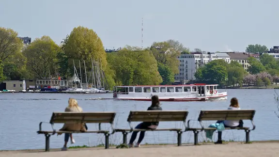 Passanten sitzen auf Bänken und genießen das sonnige Wetter an der Hamburger Außenalster. Im Hintergrund fährt ein Alsterdampfer vorbei. | picture alliance / dpa, Marcus Brandt Passanten sitzen auf Bänken und genießen das sonnige Wetter an der Hamburger Außenalster. Im Hintergrund fährt ein Alsterdampfer vorbei.