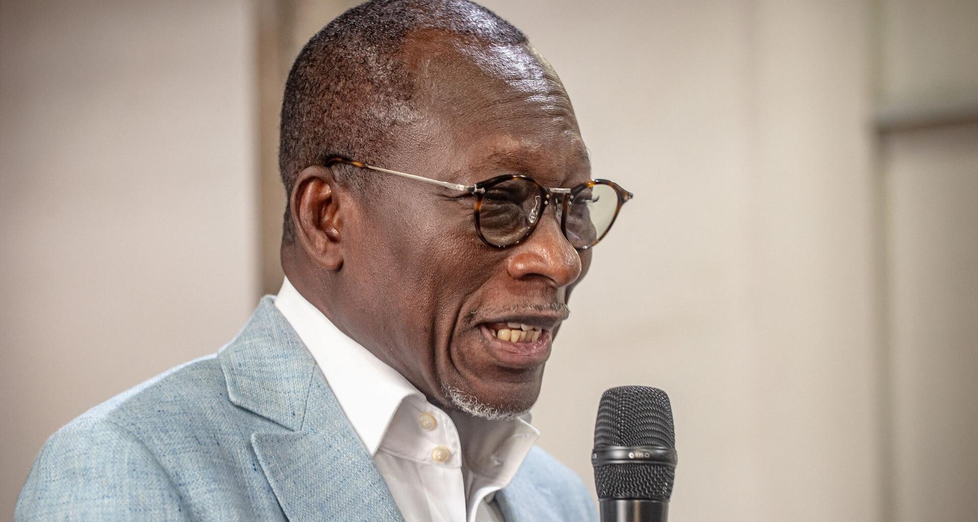 Ein schwarzer Mann mit kurzen Haaren und runder Brille spricht in ein Mikrofon. Er trägt ein weißes Hemd und ein graues Sakko.