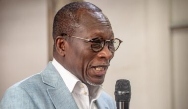 Ein schwarzer Mann mit kurzen Haaren und runder Brille spricht in ein Mikrofon. Er trägt ein weißes Hemd und ein graues Sakko.