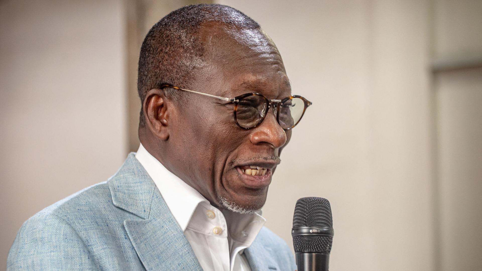 Ein schwarzer Mann mit kurzen Haaren und runder Brille spricht in ein Mikrofon. Er trägt ein weißes Hemd und ein graues Sakko.