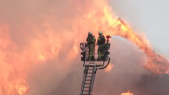 Einsatzkräfte der Feuerwehr löschen einen Brand auf einem landwirtschaftlichen Hof von einer Drehleiter aus.