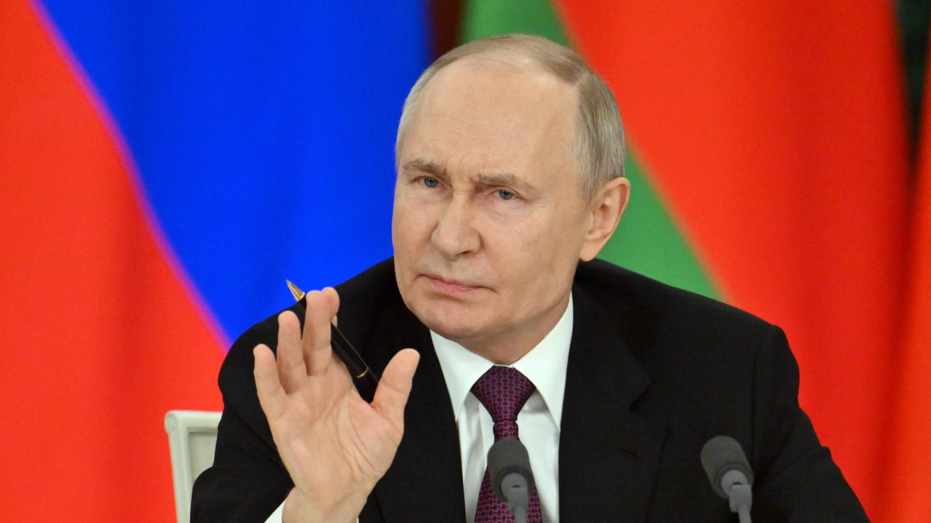 Wladimir Putin sitzt vor einer Fahne, blickt skeptisch und erhebt die rechte Hand.