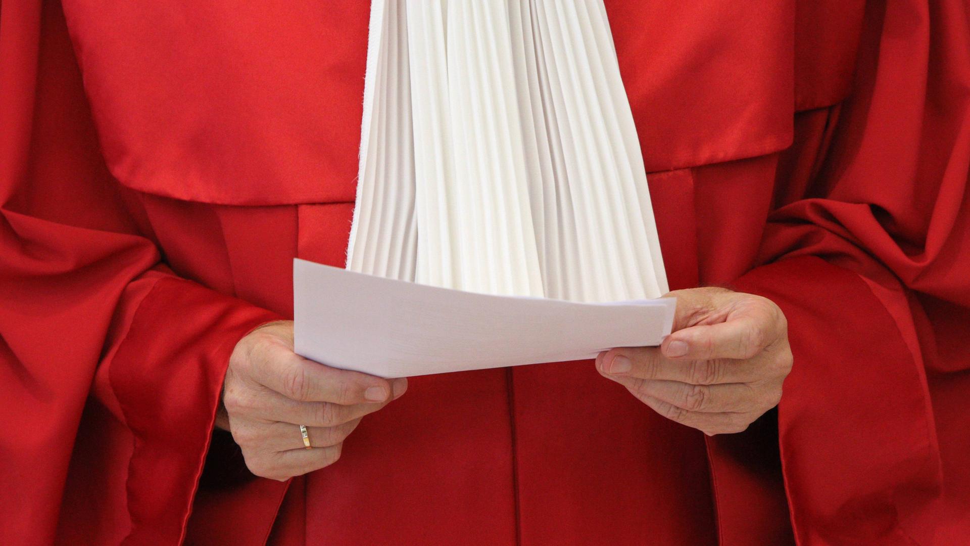 Die rote Robe eines Richters des Bundesverfassungsgerichts. Die Person hält ein Papier in der Hand. Kopf und Beine sind nicht zu sehen.