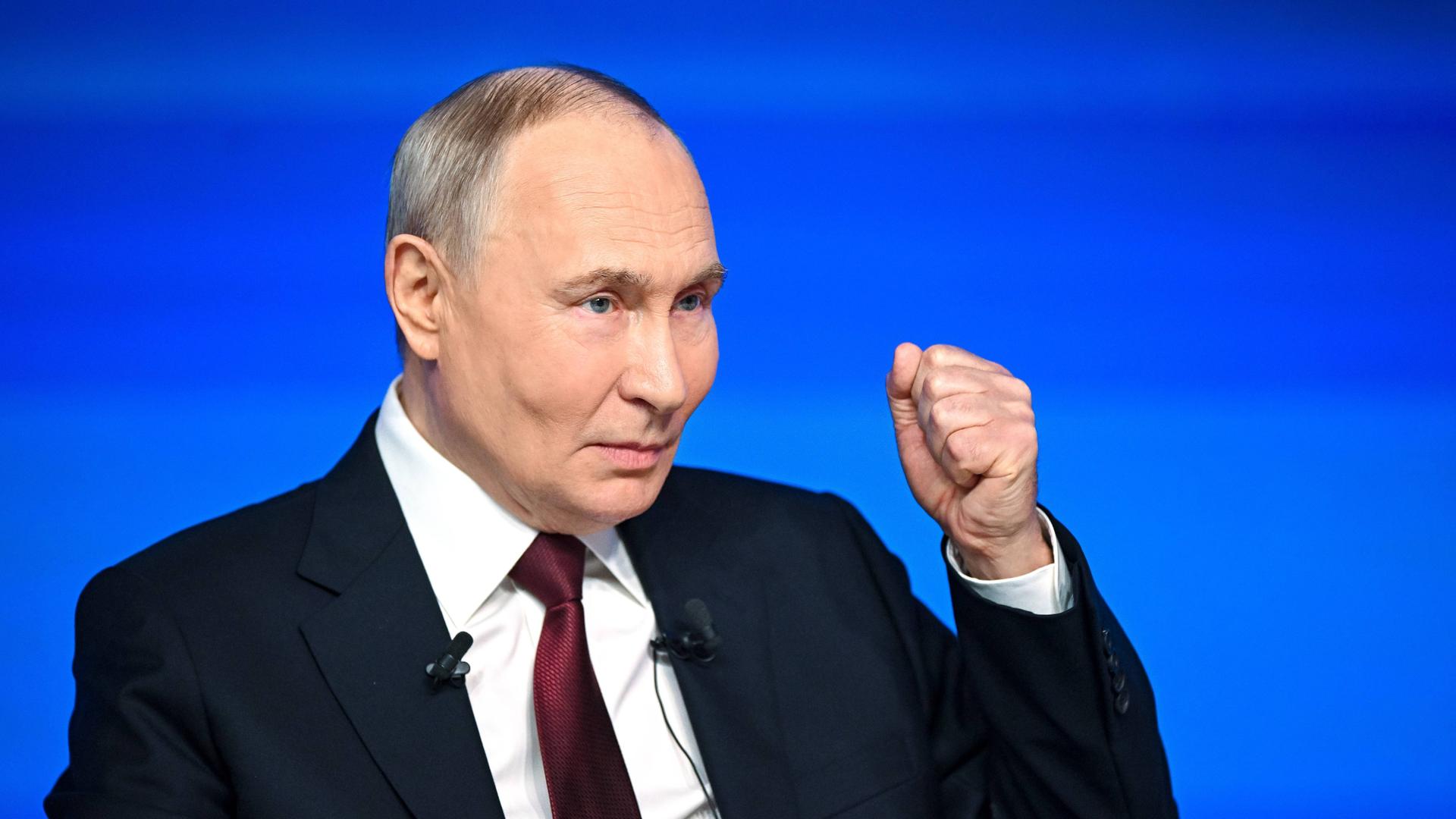 Russlands Präsident Putin steht vor einem blauen Hintergrund und hebt die linke Hand zur Faust.