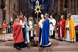 In einer großen Kirche stehen mehrere Kinder in farbigen Gewändern und mit Kronen auf dem Altarraum. Sie stellen die Sternsinger dar. Ein Kind in blauem Umhang übergibt einen großen goldenen Sternstab an andere Kinder. Im Hintergrund stehen Geistliche in weißen liturgischen Gewändern. Kerzen, Kirchenbänke und hohe Steinwände sind zu sehen. In einer großen Kirche stehen mehrere Kinder in farbigen Gewändern und mit Kronen auf dem Altarraum. Sie stellen die Sternsinger dar. Ein Kind in blauem Umhang übergibt einen großen goldenen Sternstab an andere Kinder. Im Hintergrund stehen Geistliche in weißen liturgischen Gewändern. Kerzen, Kirchenbänke und hohe Steinwände sind zu sehen.