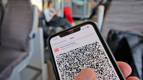 Ein Handydisplay zeigt ein Deutschlandticket an, im Hintergrund ein Bahnabteil.