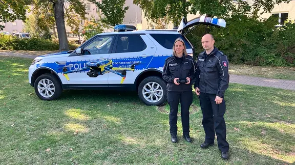 Zwei Einsatzkräfte der Polizei stehen vor einem Polizeiauto und starten eine Drohne.
