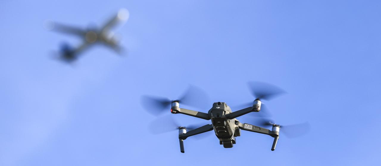 Eine Drohne fliegt am Himmel, im Hintergrund verschwommen ein Flugzeug. | dpa Eine Drohne fliegt am Himmel, im Hintergrund verschwommen ein Flugzeug.