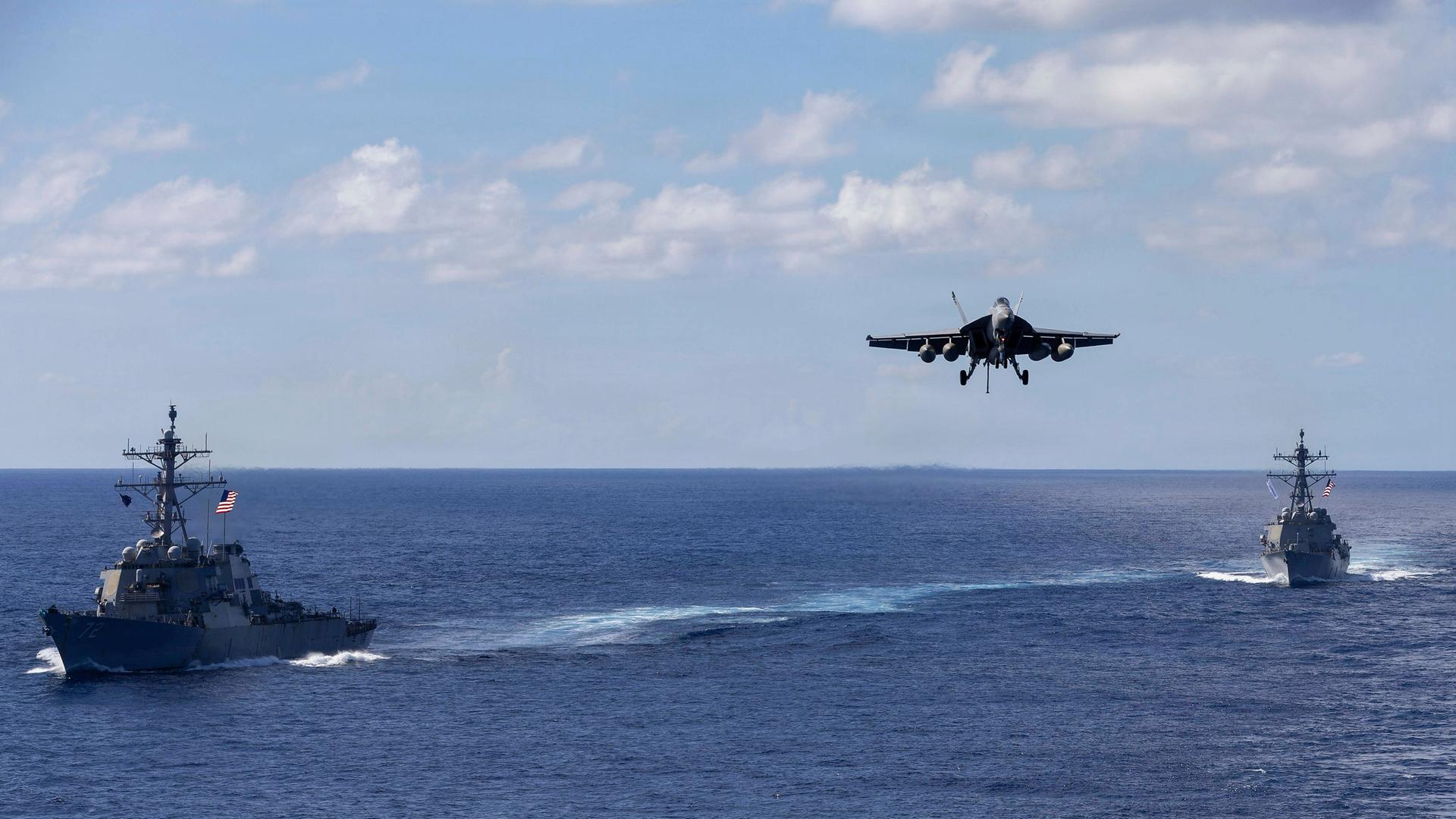 Zwei militärische Schiffe fahren auf dem offenen Meer, dazwischen setzt ein Kampfflugzeug zum Landeanflug an.