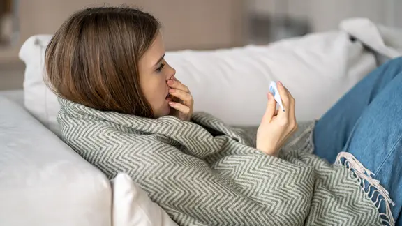 Eine hustende Frau schaut auf ein Fieberthermometer.