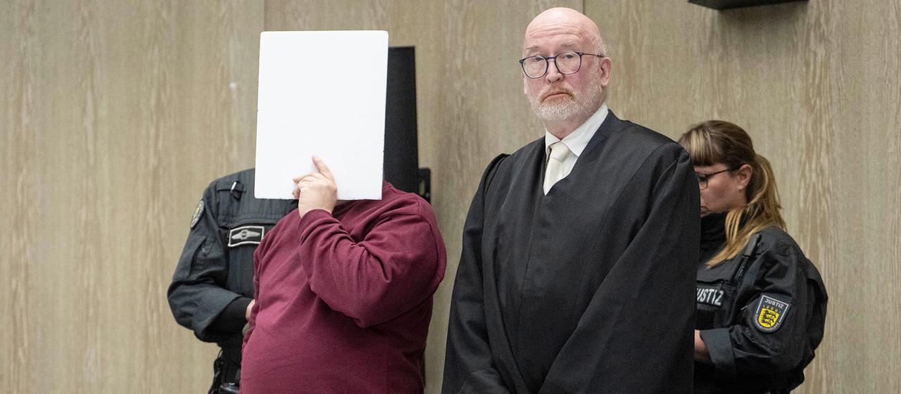 Der Angeklagte steht mit einem Aktendeckel vor seinem Gesicht im Gerichtssaal. | dpa Der Angeklagte steht mit einem Aktendeckel vor seinem Gesicht im Gerichtssaal.