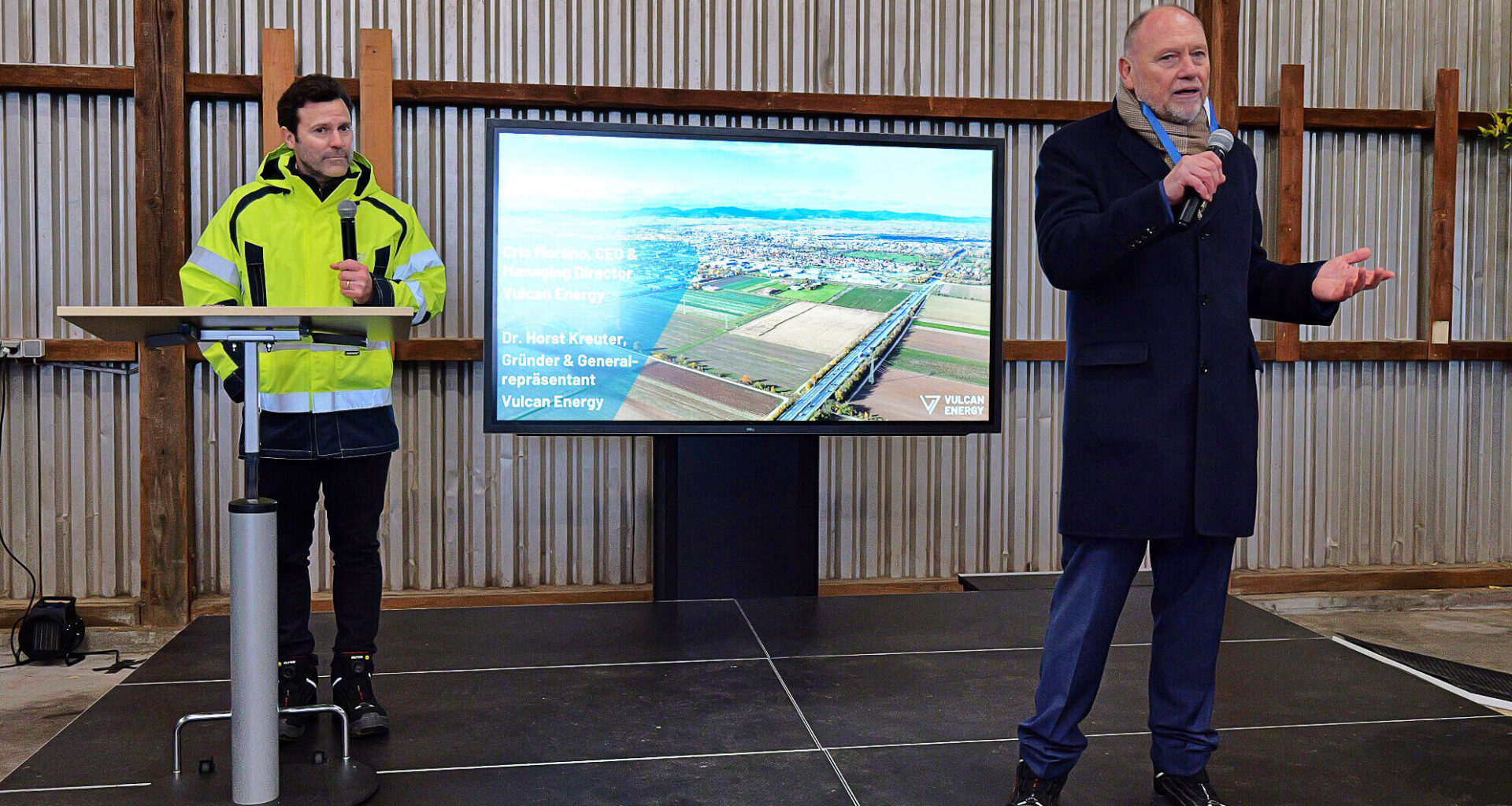 Zwei Männer mit Mikrofonen auf einer Bühne, Bildschirm mit Text "Vulcan Energy".