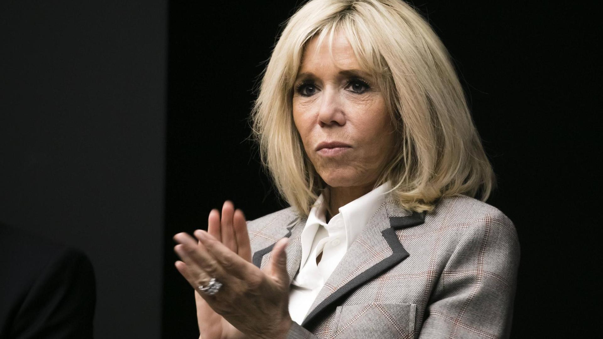 Macron im grauen Kostüm steht vor einem dunklen Hintergrund und klatscht in die Hände. 