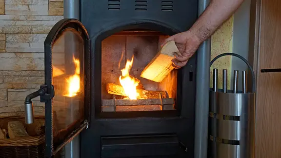 Ein Stück Holz wird in den brennenden Kamin gelegt.