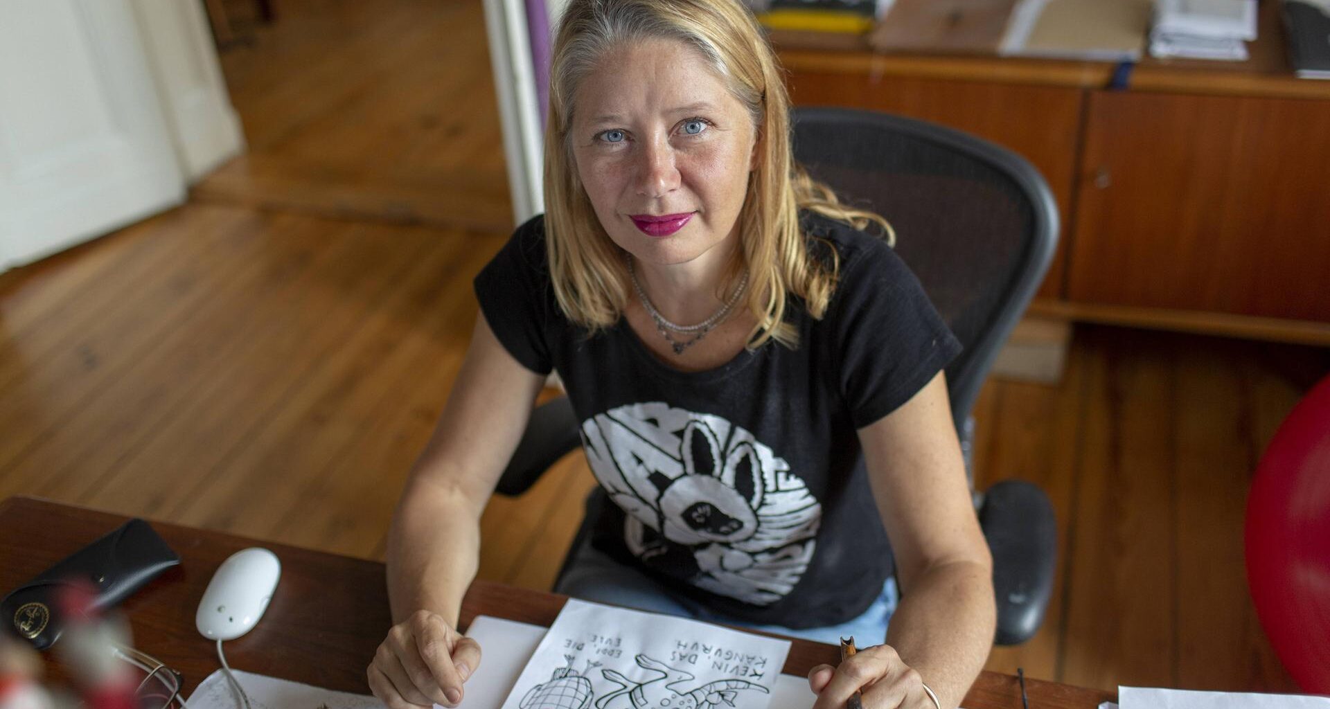 Kat Menschik sitzt an ihrem Schreibtisch. Sie hat blondes, schulterlanges Haar und trägt roten Lippenstift. Vor ihr liegt eine Zeichung in Schwarzweiß.