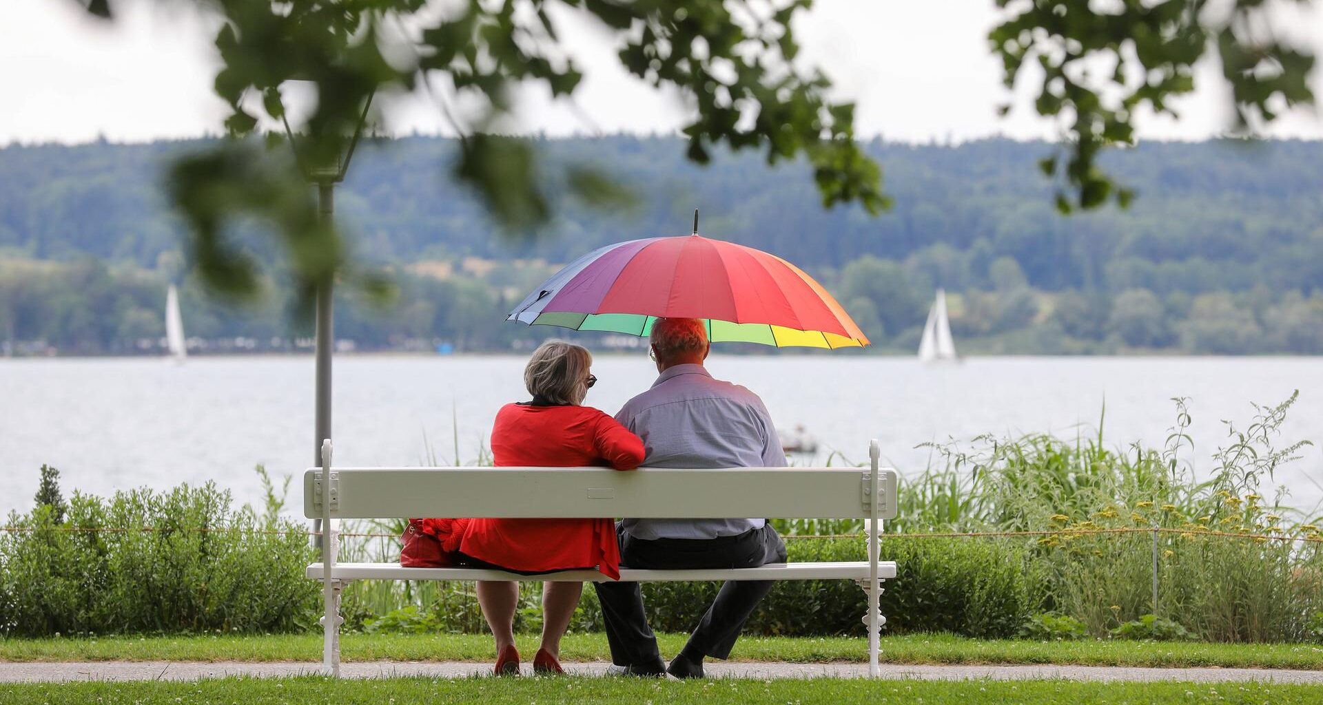 Ein Mann und eine Frau sitzen am Ufer des Bodensees mit einem Sonnenschirm in der Hand auf einer Bank.