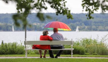 Ein Mann und eine Frau sitzen am Ufer des Bodensees mit einem Sonnenschirm in der Hand auf einer Bank.