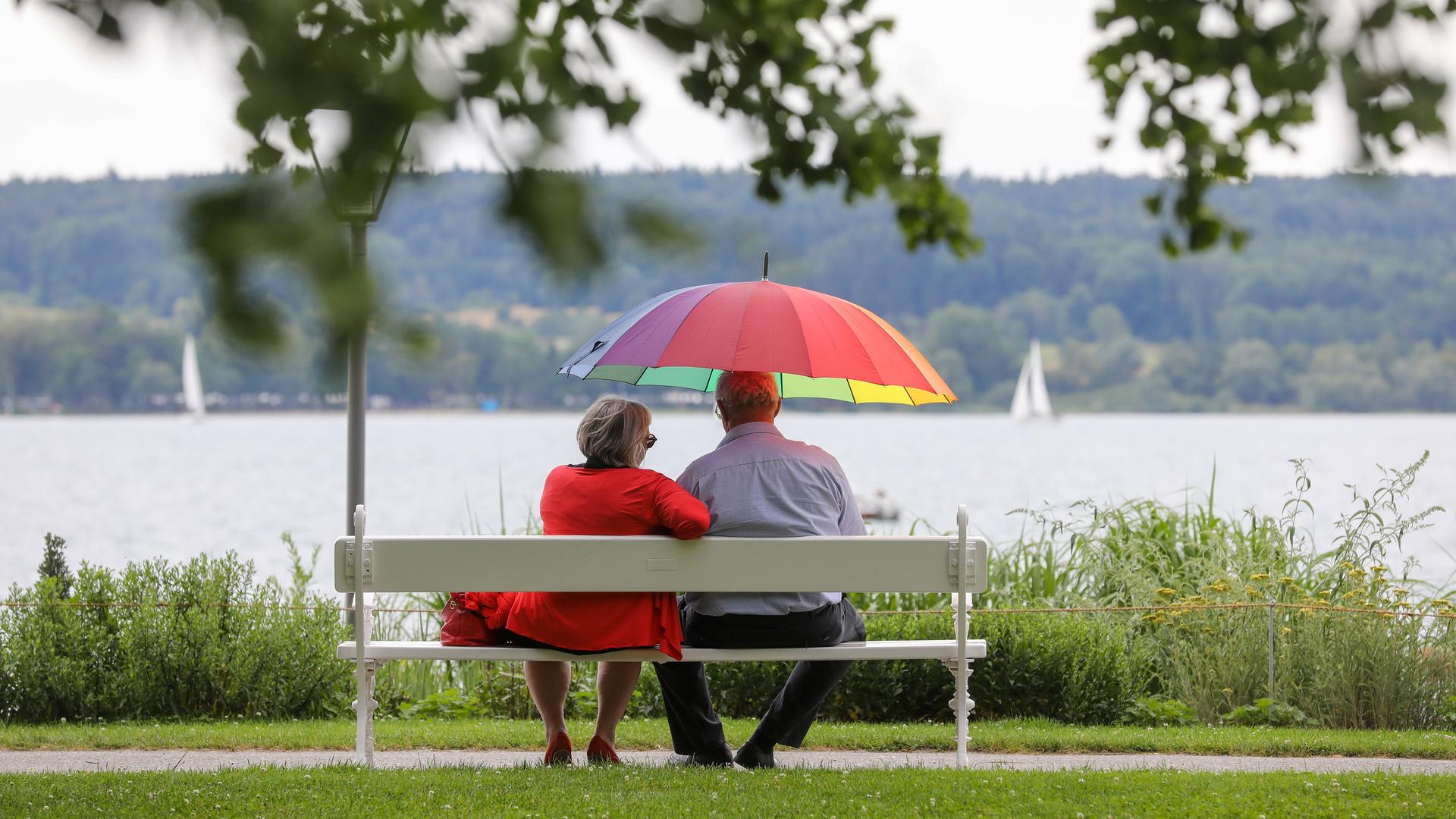 Ein Mann und eine Frau sitzen am Ufer des Bodensees mit einem Sonnenschirm in der Hand auf einer Bank. Ein Mann und eine Frau sitzen am Ufer des Bodensees mit einem Sonnenschirm in der Hand auf einer Bank.