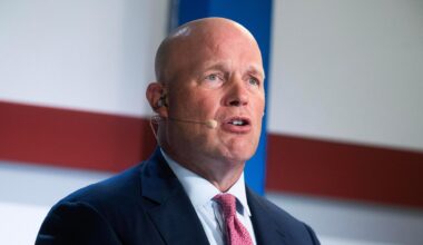 Der glatzköpfige Whitaker mit dunklem Jackett und roter Krawatte sagt etwas. Dahinter eine weiße Wand mit einem roten Quer- und einem blauen Längstreifen, die sich kreuzen.